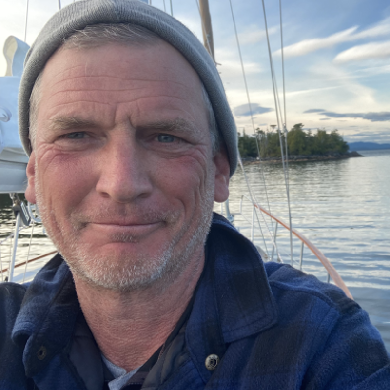 The image shows a man on a boat. He is wearing a gray beanie and a blue plaid shirt. The background includes water, trees, and a cloudy sky. The man is smiling at the camera. It seems like a peaceful day on the water.